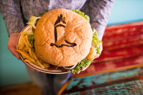 A person holds a large burger with a unique design on the bun, accompanied by a side of fries on a plate.