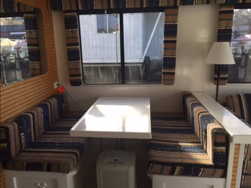 Cozy interior of a camper with a table and striped seating by a window, featuring a small heater below.
