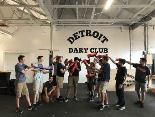 A group of young adults in a dart club playfully aim toy blasters at each other in a spacious indoor setting.