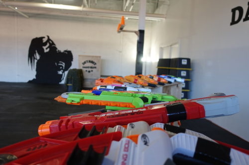 A row of colorful toy blasters lined up on a table, with a shadowy figure painted on the wall in the background.