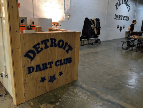 Interior of the Detroit Dart Club, featuring a wooden counter and a person at a table in a spacious, industrial setting.