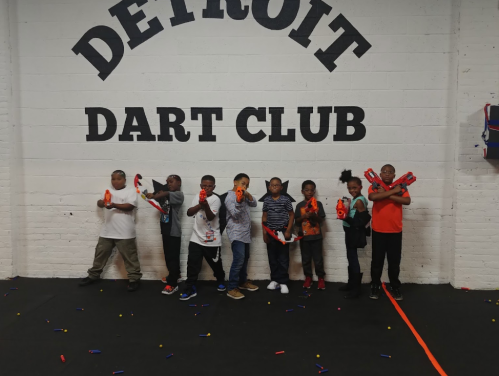 A group of children holding toy dart guns, standing in front of a wall that says "Detroit Dart Club."