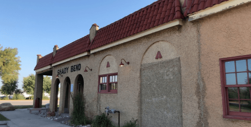 A building with a red-tiled roof and textured beige walls, featuring arched windows and the sign "Shady Bend."