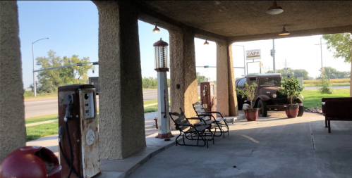 Vintage gas station with old pumps, chairs, and a classic car under a covered area, surrounded by greenery.