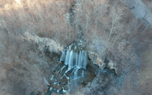 Aerial view of a waterfall cascading down rocky cliffs, surrounded by bare trees and a winding road nearby.