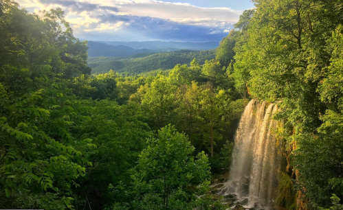 A scenic view of a waterfall cascading through lush green trees, with mountains in the background under a cloudy sky.