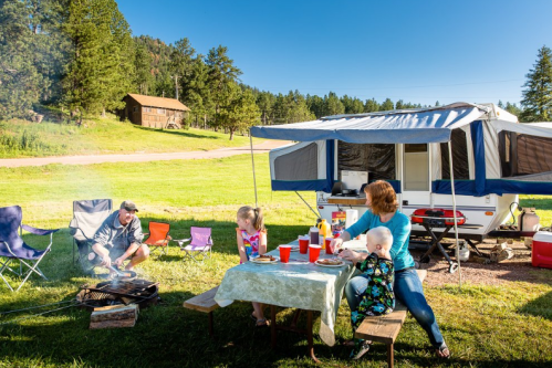 A family enjoys a camping meal outdoors, with a grill, picnic table, and a camper in a scenic, green setting.