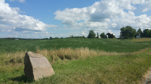 A grassy field under a blue sky with clouds, featuring a stone marker and a distant farmhouse.