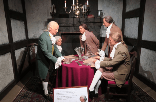 Wax figures of historical figures gathered around a table in a dimly lit room, discussing documents and plans.