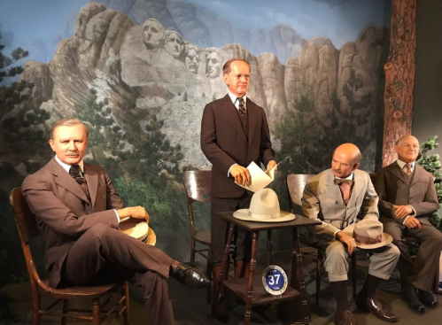 Four lifelike wax figures in vintage attire, seated and standing, with a mountain backdrop resembling Mount Rushmore.