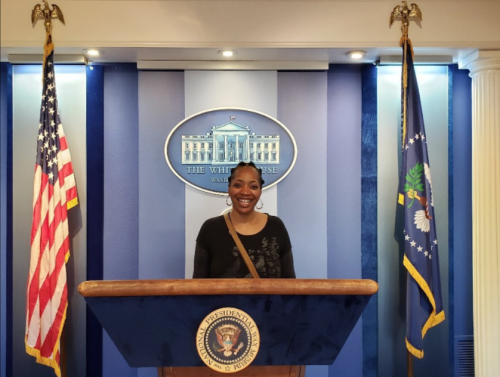 A smiling person stands at a podium in the White House press room, flanked by American flags.
