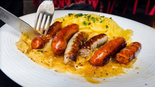 A plate of assorted sausages served with sauerkraut and garnished with herbs. A fork and knife are placed beside the dish.