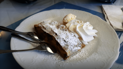 A slice of apple strudel dusted with powdered sugar, served with whipped cream and a small cookie on a white plate.