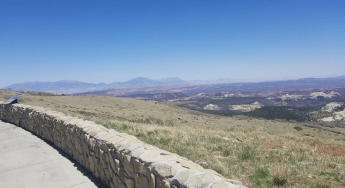 A scenic view of rolling hills and distant mountains under a clear blue sky, with a stone wall in the foreground.
