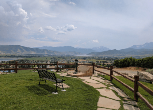 Scenic view of mountains and a valley from a park, featuring a bench and stone pathway under a cloudy sky.