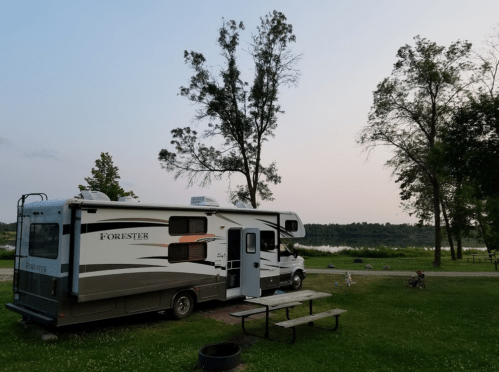 An RV parked by a serene lake at dusk, surrounded by trees and a picnic table on the grass.