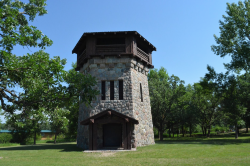 A stone tower with a wooden roof surrounded by trees and green grass under a clear blue sky.
