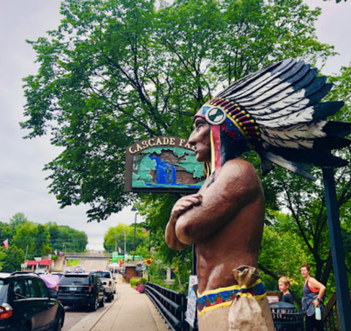 A large Native American statue stands near a sign for Cascade Falls, surrounded by trees and parked cars.
