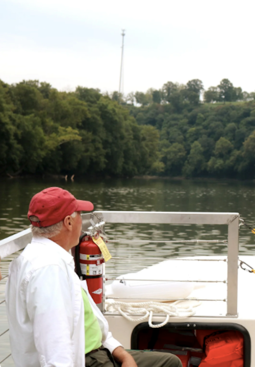 A man in a red cap sits on a boat, gazing at the calm water and lush green trees in the background.