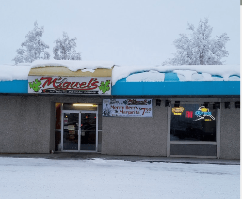 A snowy scene of Miguel's Authentic Mexican Cuisine restaurant, featuring a colorful sign and neon lights.