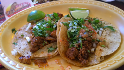 Two tacos filled with seasoned meat, topped with onions, cilantro, and salsa, served with lime wedges on a yellow plate.