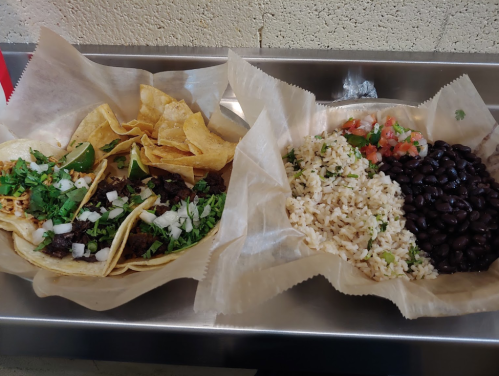 A platter featuring tacos with cilantro and onions, alongside rice, black beans, and pico de gallo, all in paper trays.