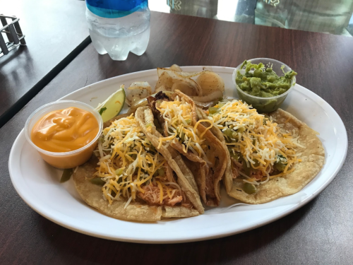 A plate of three tacos topped with cheese, served with guacamole, a side of potatoes, and a cup of orange sauce.