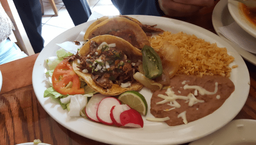 A plate of tacos with meat, onions, and cilantro, served with rice, refried beans, and fresh vegetables.
