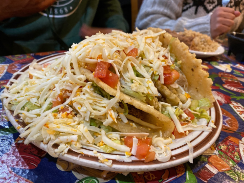 A large plate of tacos piled high with lettuce, tomatoes, and shredded cheese on a colorful tablecloth.