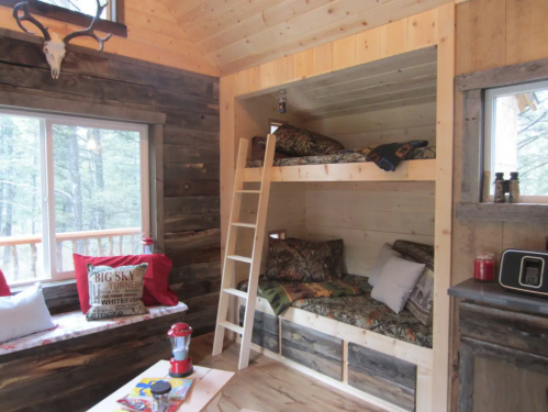 Cozy cabin interior featuring a bunk bed, wooden walls, and a window with forest views. Decor includes cushions and a lantern.