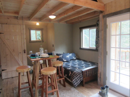 Cozy cabin interior with a bed, wooden stools, and a dining table near large windows and a door to the outside.
