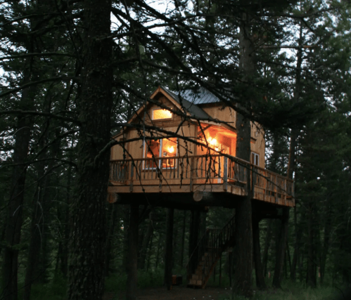 A cozy treehouse illuminated at dusk, nestled among tall trees with a wooden deck and stairs leading down.