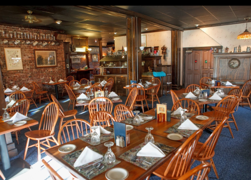 Cozy restaurant interior with wooden tables, chairs, and neatly set diningware, featuring brick walls and warm lighting.