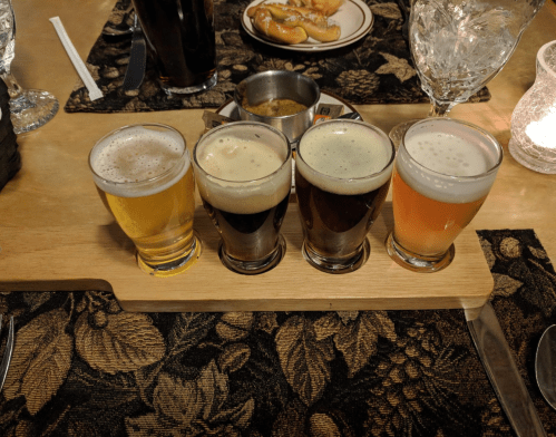A wooden flight board with four small glasses of different beers, set on a floral-patterned tablecloth.