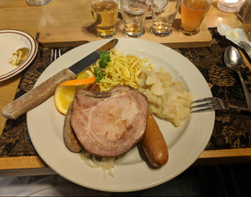 A plate of food featuring sliced meat, sausage, noodles, and a side of applesauce, garnished with orange slices.
