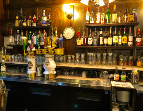 A wooden bar with shelves of various liquor bottles, a clock, and a row of clean glasses on the counter.