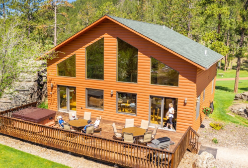 A large log cabin with large windows, surrounded by trees, featuring a deck with outdoor seating and a hot tub.