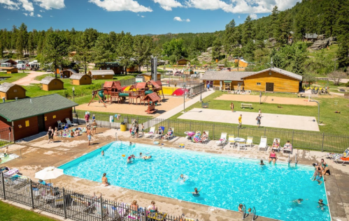 A sunny outdoor pool area with people swimming, surrounded by cabins, playgrounds, and green trees in the background.