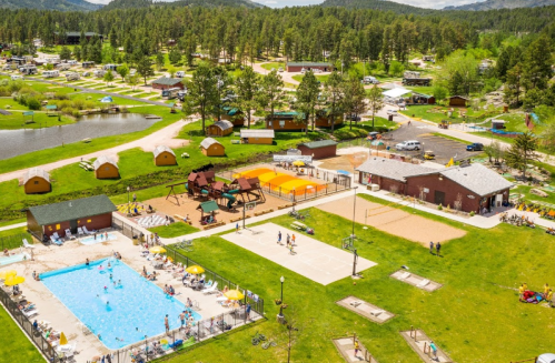 Aerial view of a campground featuring a pool, playground, sports courts, and cabins surrounded by trees.