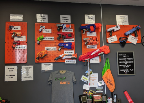 A wall display of various toy dart blasters, with pricing and a shirt for sale at a dart-themed venue.