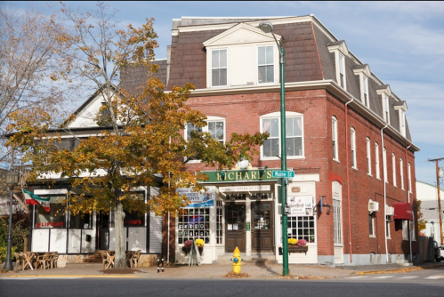 A brick building with a green awning, featuring a restaurant and shops, surrounded by autumn trees.