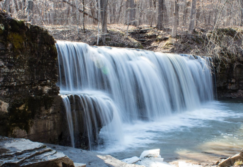 A serene waterfall cascades over rocky ledges, surrounded by bare trees and a tranquil pool of water below.