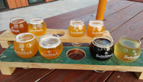 A wooden flight holder with eight glasses of various craft beers on a table.