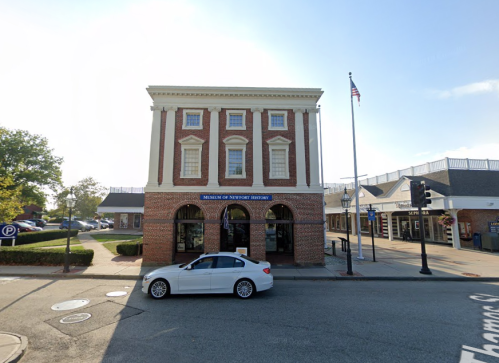 A brick building with large windows, labeled "Museum of Newport History," beside a street and parked cars.