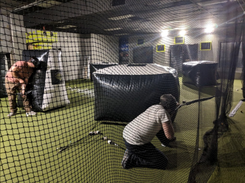 Two players crouch behind inflatable barriers in an indoor paintball arena, with bright lights overhead.
