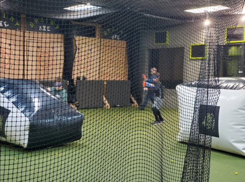 A person plays in an indoor paintball arena, surrounded by inflatable barriers and netting.