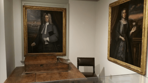 Two portraits hang on the wall, one of a man and one of a woman, beside a wooden box and a chair in a museum setting.