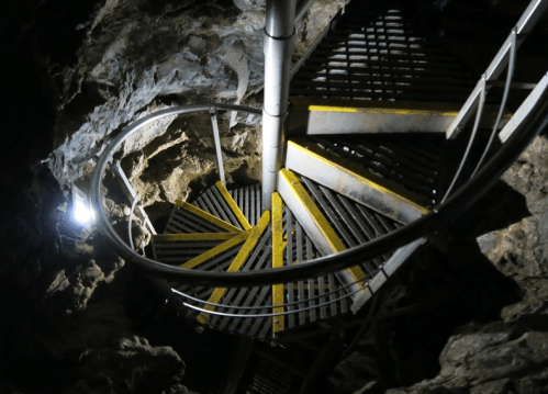 A spiral staircase descends into a dark cave, illuminated by a single light, with rocky walls surrounding it.