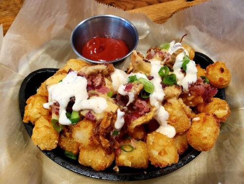 A sizzling platter of loaded tater tots topped with bacon, green onions, and drizzled with sauce, served with ketchup.