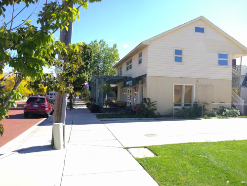A two-story yellow building with a porch, surrounded by greenery, on a sunny street with parked cars.